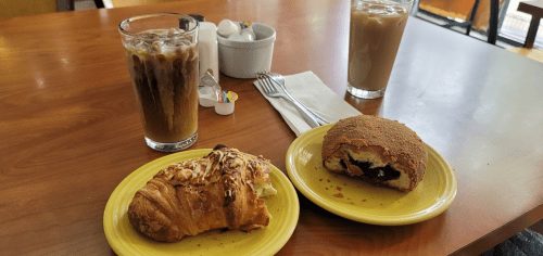 A table with two plates: one with a croissant and one with a pastry, alongside two glasses of iced coffee.