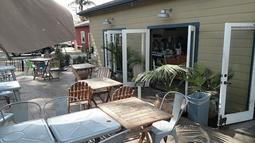 Outdoor seating area of a café with wooden tables, chairs, and potted plants, leading to an entrance.