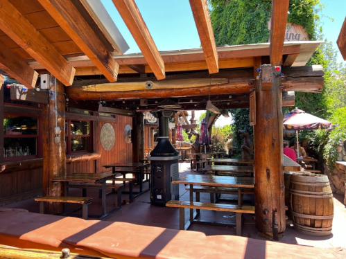 Outdoor dining area with wooden beams, benches, and a central heater, surrounded by greenery and rustic decor.