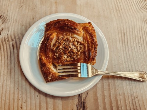 A flaky pastry on a white plate, topped with nuts, accompanied by a fork, resting on a wooden table.