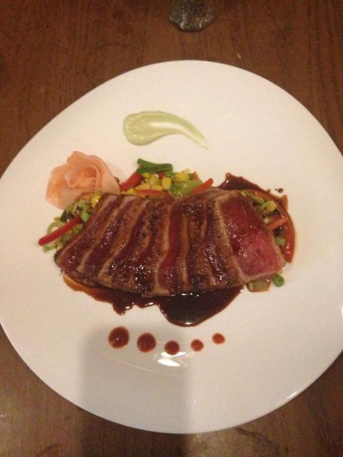 A plate of seared steak slices with colorful vegetables and a drizzle of sauce, garnished with pickled ginger and a green sauce.