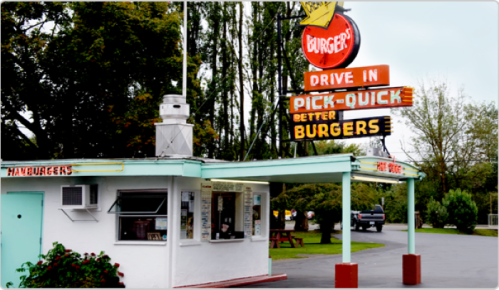 A retro drive-in burger stand with a colorful sign, surrounded by trees and a parking area.