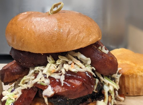 A close-up of a hearty burger topped with coleslaw and sausage, served on a toasted bun with a side of cornbread.