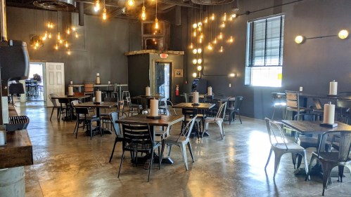 A modern dining area with wooden tables, metal chairs, and warm pendant lights against a dark wall.