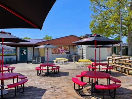 Outdoor seating area with red picnic tables and umbrellas at Pizza King, surrounded by trees and a clear blue sky.