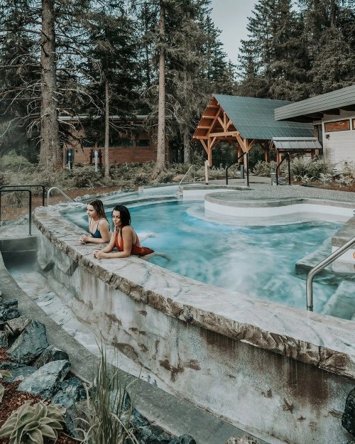 Two women relax in a hot tub surrounded by trees and a cozy cabin in a serene outdoor setting.