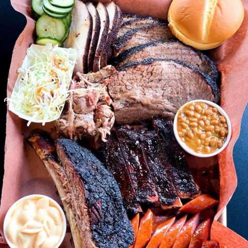 A barbecue platter featuring brisket, pulled pork, ribs, sausage, coleslaw, pickles, baked beans, and a bun.