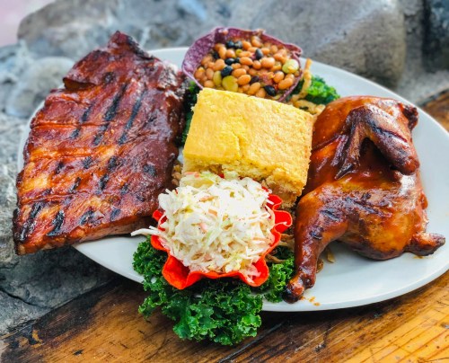 A plate of grilled ribs, roasted chicken, cornbread, coleslaw, and black bean salad, garnished with kale.