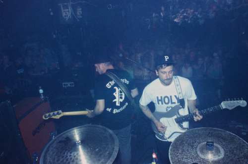 Two musicians perform on stage, one playing guitar and the other on bass, with a crowd visible in the background.