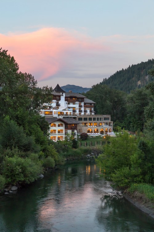 A scenic view of a charming hotel by a river, surrounded by lush trees and mountains at sunset.
