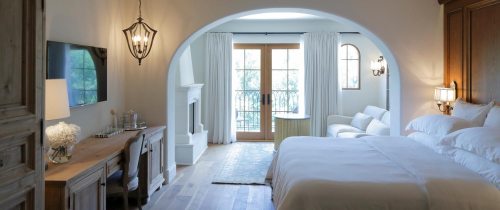Bright, airy hotel room with a large bed, desk, and balcony doors, featuring soft furnishings and natural light.