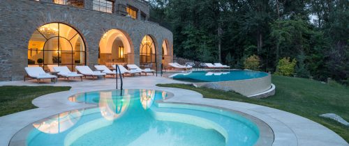 A luxurious outdoor area featuring two pools, lounge chairs, and a stone house surrounded by lush greenery.
