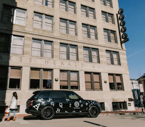 A black SUV with white letters parked outside the Press Hotel, a brick building with large windows.