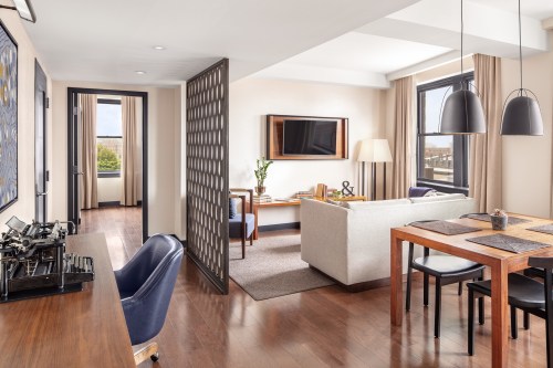 A modern hotel suite featuring a living area, dining table, and workspace with stylish decor and large windows.