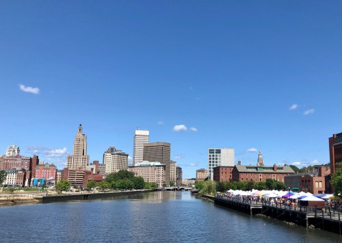 A city skyline with tall buildings along a river, featuring a blue sky and a waterfront market with tents.