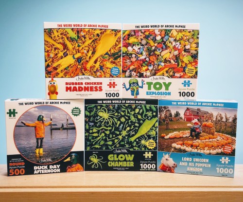 A collection of colorful jigsaw puzzle boxes featuring whimsical themes and characters from Archie McPhee.