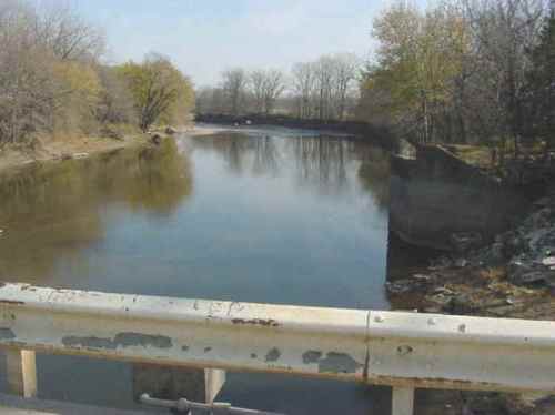 A calm river flows between trees, with a concrete barrier visible in the foreground under a clear sky.