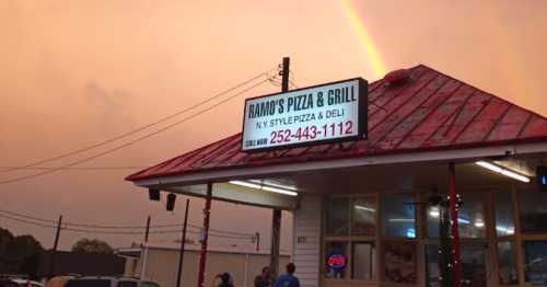 Sign for Ramo's Pizza & Grill under a colorful sky with a rainbow, featuring a red roof and outdoor seating.