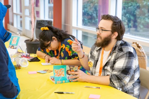 A man with a beard talks at a table with a woman writing, both surrounded by games and colorful decorations.