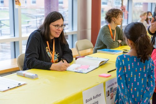 A woman with glasses signs a book for a young girl at a table during a book signing event.