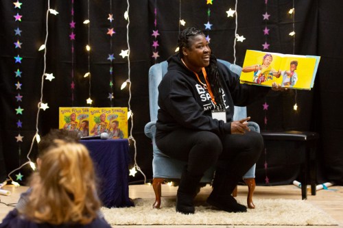A storyteller in a black hoodie sits in a chair, holding a colorful book, with children listening attentively.