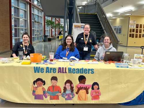 Four women smile behind a table with a banner reading "We Are ALL Readers," in a bright, open space.