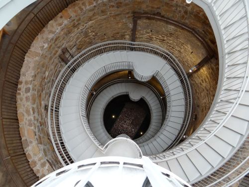 A spiral staircase viewed from above, featuring stone walls and metal railings, leading down into a dark center.