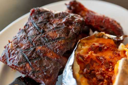 A plate featuring grilled meat, charred on the outside, alongside a baked potato topped with bacon bits.