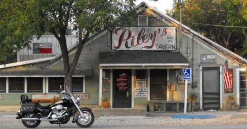 A vintage tavern named Riley's with a motorcycle parked outside, surrounded by trees and an American flag.