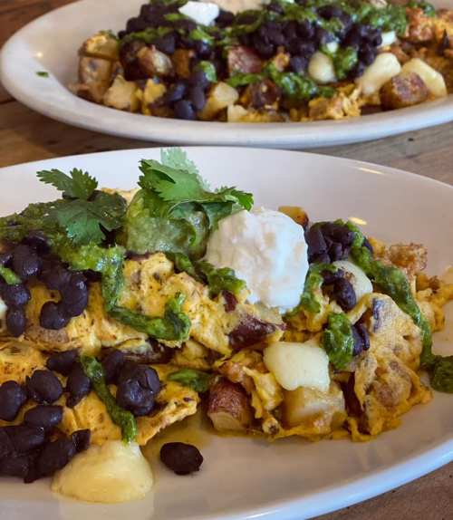 A plate of loaded breakfast nachos topped with eggs, black beans, green sauce, sour cream, and cilantro.