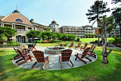 A scenic outdoor area with wooden chairs arranged around a fire pit, surrounded by lush greenery and a hotel in the background.