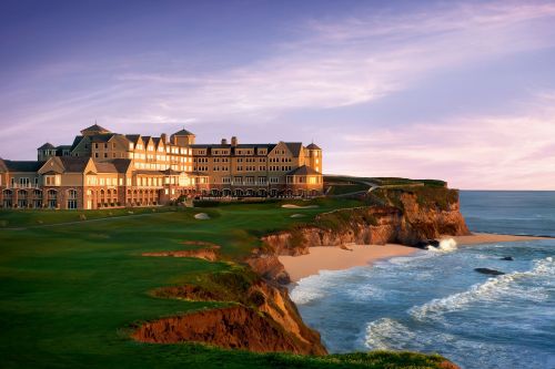 A luxurious hotel perched on a cliff overlooking the ocean, with lush green lawns and a sunset sky in the background.