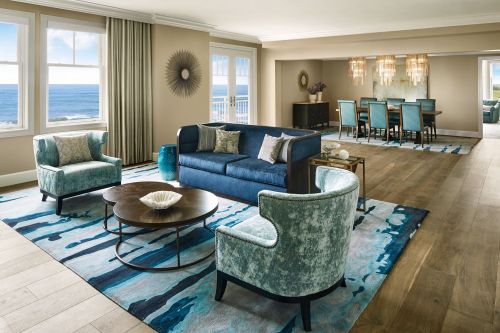 A modern living room with blue furniture, a round coffee table, and ocean views through large windows.