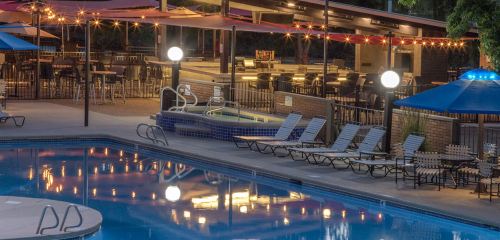 A serene pool area at night, featuring lounge chairs, umbrellas, and a bar with warm lighting.