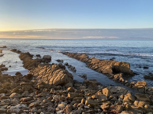 Rocky shoreline at sunset, with calm ocean waves and a clear sky in the background.