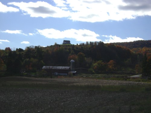 A scenic view of a farm with a barn and silo, set against a backdrop of colorful autumn trees and a cloudy sky.