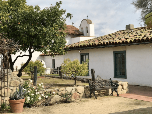 A serene courtyard with a stone bench, orange tree, and white buildings under a cloudy sky.
