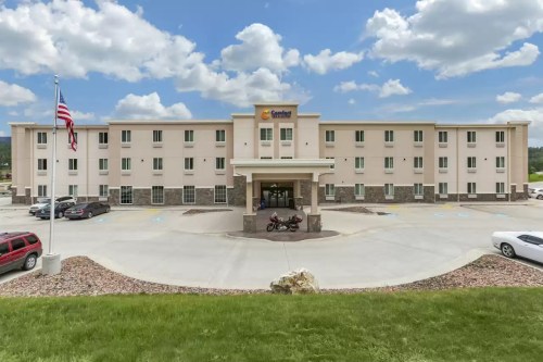 Exterior view of a three-story hotel with a central entrance, surrounded by parking and green grass under a cloudy sky.