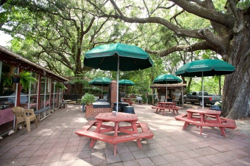 Outdoor seating area with red picnic tables and green umbrellas under large trees, creating a shaded, inviting atmosphere.