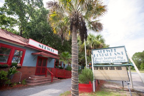 A seafood restaurant with a red roof and palm trees, featuring a sign that reads "Stop Here for Good Food."