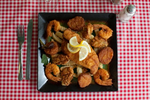 A black plate filled with fried fish, shrimp, and hushpuppies, garnished with lemon slices, on a red checkered tablecloth.