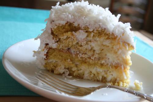 A slice of coconut cake topped with shredded coconut on a white plate, with a fork beside it.