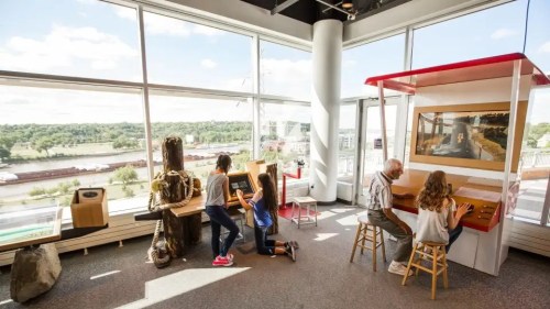 Children and an elderly man interact with exhibits in a bright, spacious room with large windows and a scenic view.