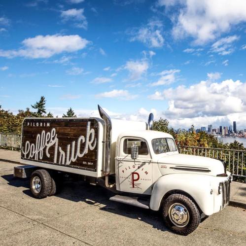 A vintage coffee truck parked with a city skyline and blue sky in the background. The truck features a wooden sign.