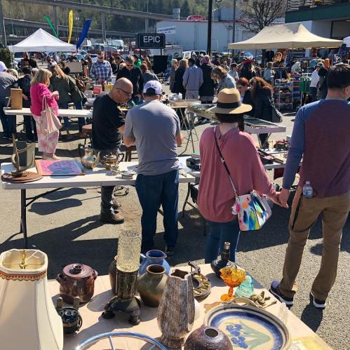 A bustling outdoor market with people browsing tables filled with various items and antiques under a sunny sky.