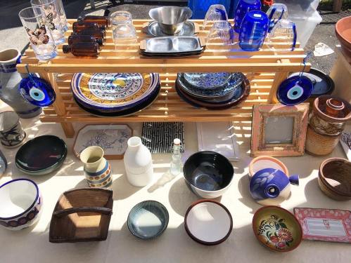 A display of various colorful dishes, bowls, and glassware arranged on a wooden shelf at an outdoor market.