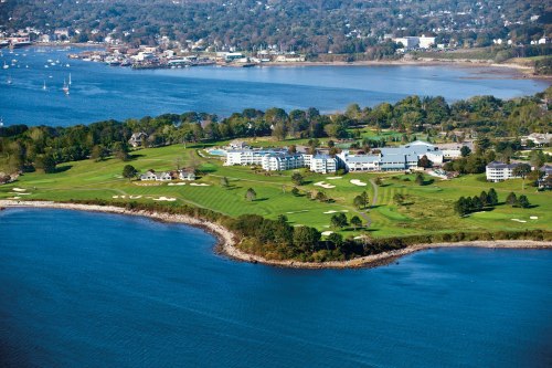 Aerial view of a coastal golf course with lush greens, a hotel, and a marina in the background.