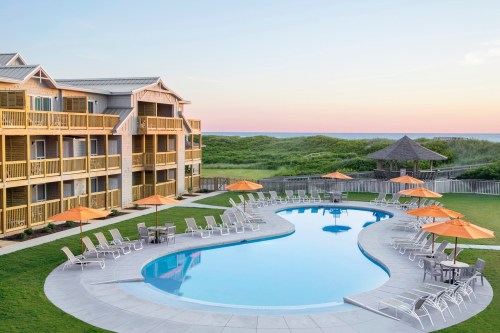 A serene hotel pool area with orange umbrellas, lounge chairs, and ocean views at sunset.