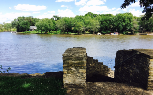A serene river scene with lush green trees, a house in the background, and stone steps leading down to the water.