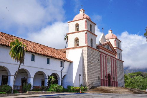 Historic church with pink domes, stone facade, and palm trees, set against a blue sky with clouds.